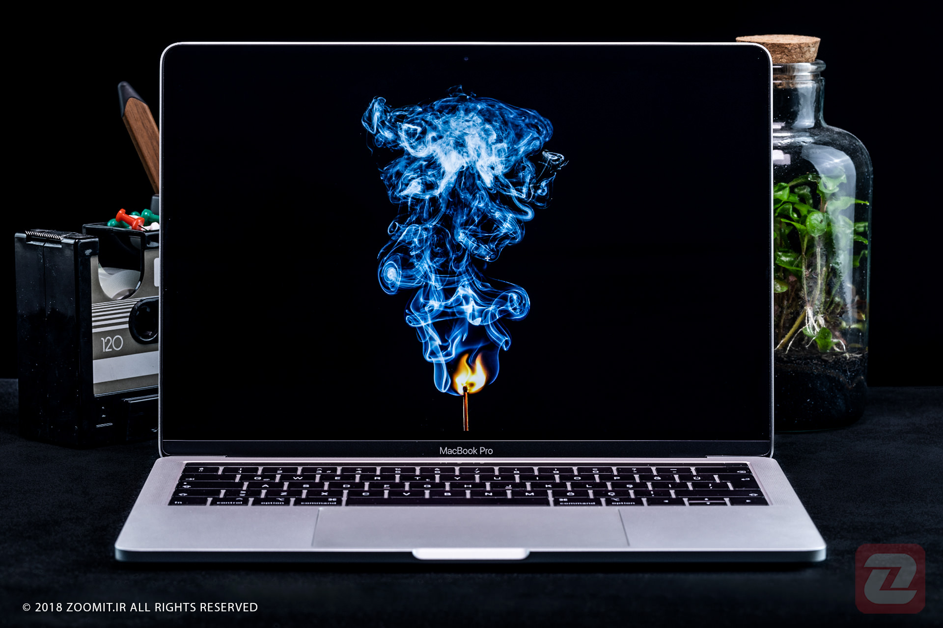 تصویری از یک مامپیوتر در کنار یک رادیو و گلدان شیشه ای و همینطور نمایشگر کامپیوتر در حال نشان دادن یک شعله با دود ابی است 